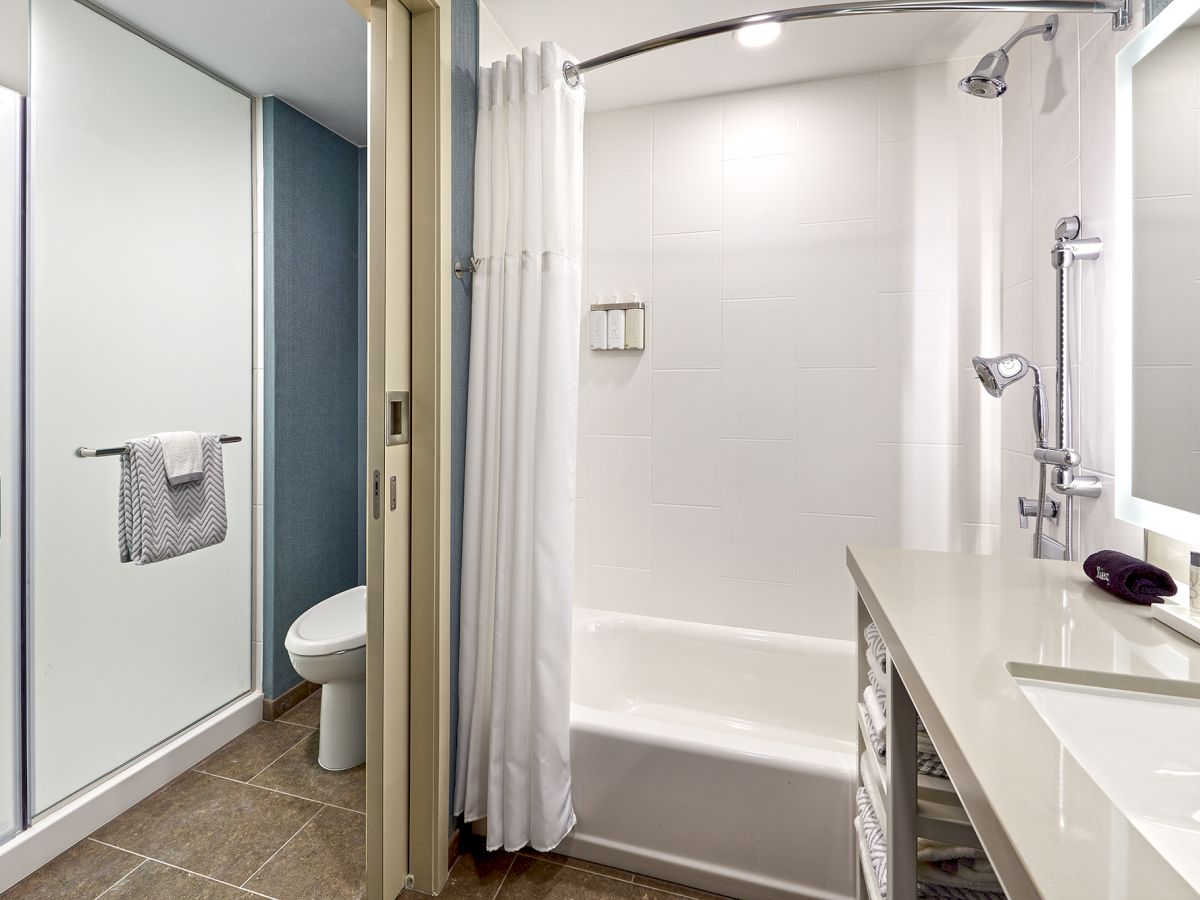 This image shows a modern bathroom with a shower-over-tub setup, toilet, sink, and mirror, featuring white and neutral tones and plenty of natural light.