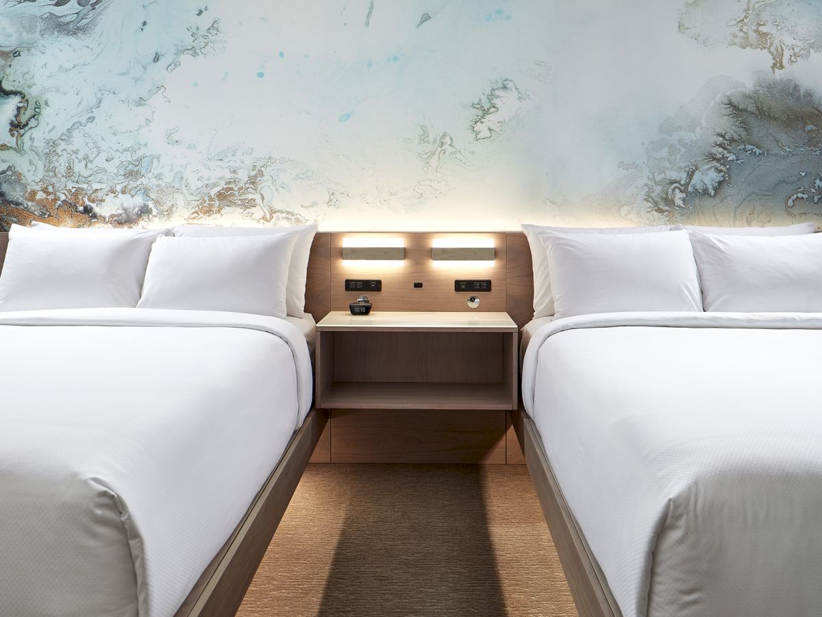 Two neatly made beds with white linens are adjacent to each other. They share a wooden nightstand with built-in lights and outlets on the wall.