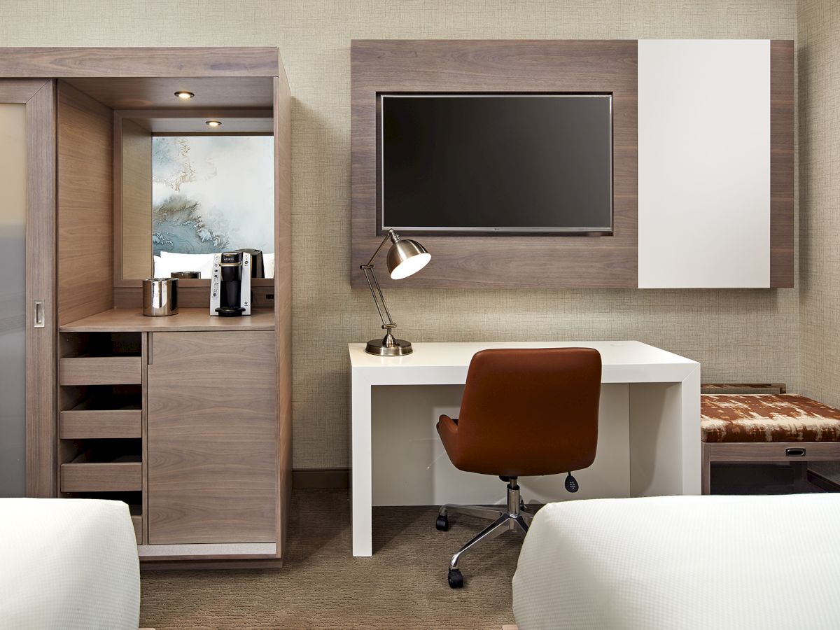 A modern hotel room features a TV, desk with lamp and chair, a cabinet with drink amenities, and a bench. Two beds are partially visible.