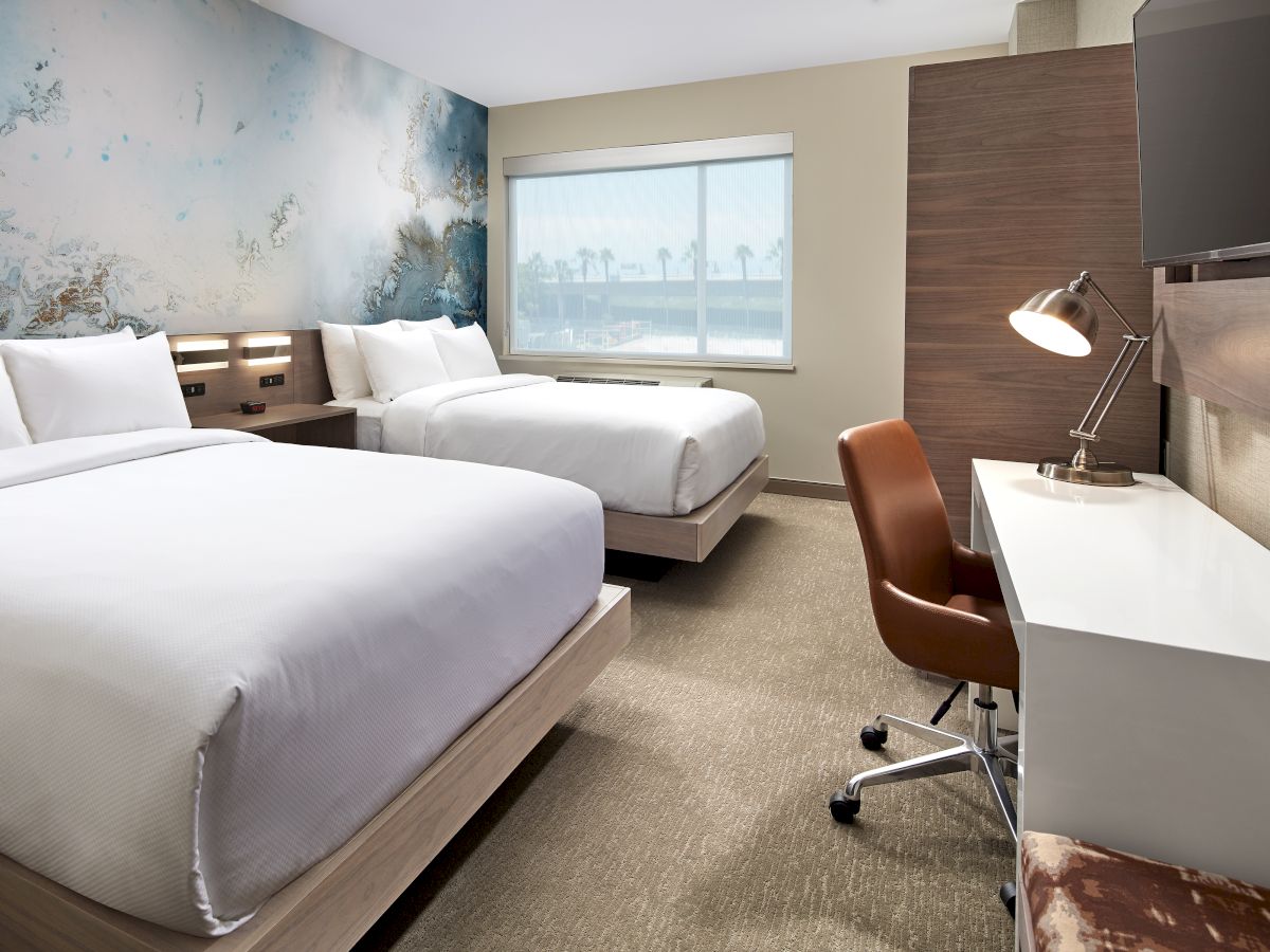 A modern hotel room has two beds, a large window, a desk with a lamp, a chair, and a flat-screen TV mounted on the wall.