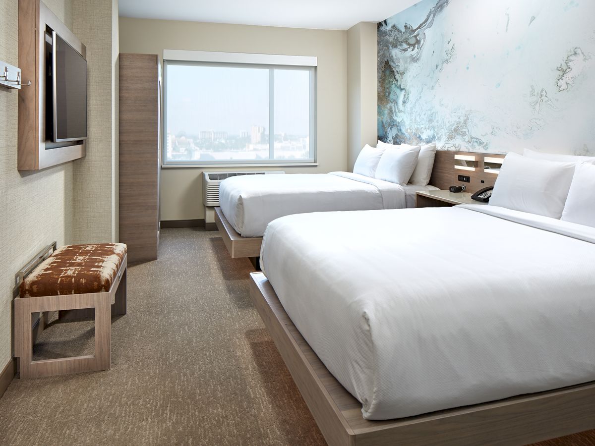 A modern hotel room features two neatly made beds, a wall-mounted TV, a window with a view, a bench, and minimalistic decor with an abstract wall art.