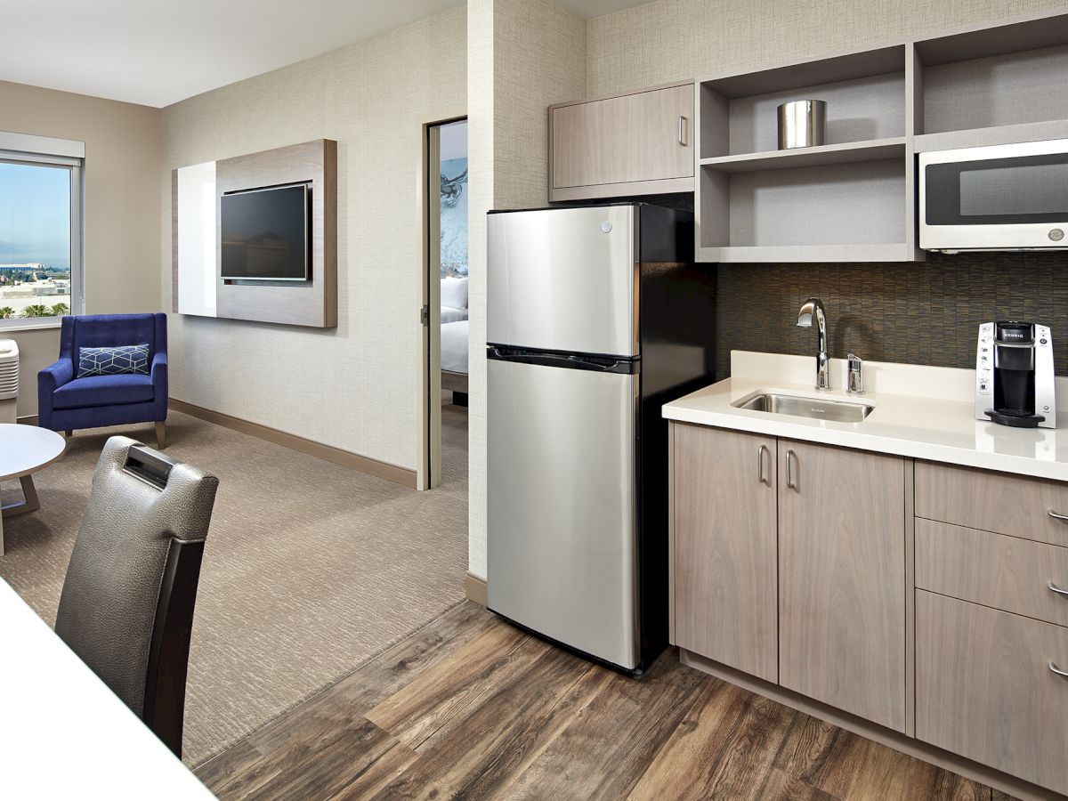 This image shows a modern hotel room with a kitchenette, including a fridge, microwave, sink, and coffee maker, and a seating area with a view.