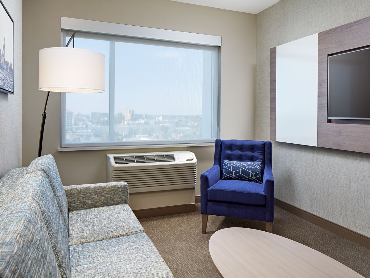 A modern living room with a blue chair, grey sofa, a wall-mounted TV, a large window, and a floor lamp next to an air conditioning unit.