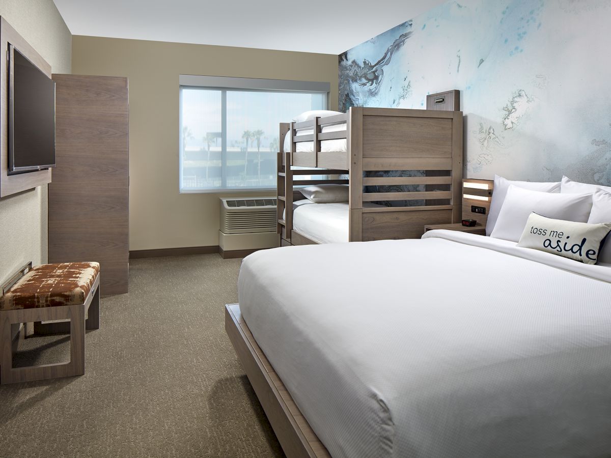 The image shows a modern hotel room with a double bed, bunk beds, a flat-screen TV, a bench, and a window with a view, decorated in neutral tones.