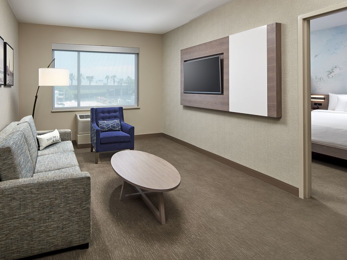 The image shows a modern hotel room with a gray sofa, blue chair, oval coffee table, mounted TV, window with a view, and an adjacent bedroom with a bed.