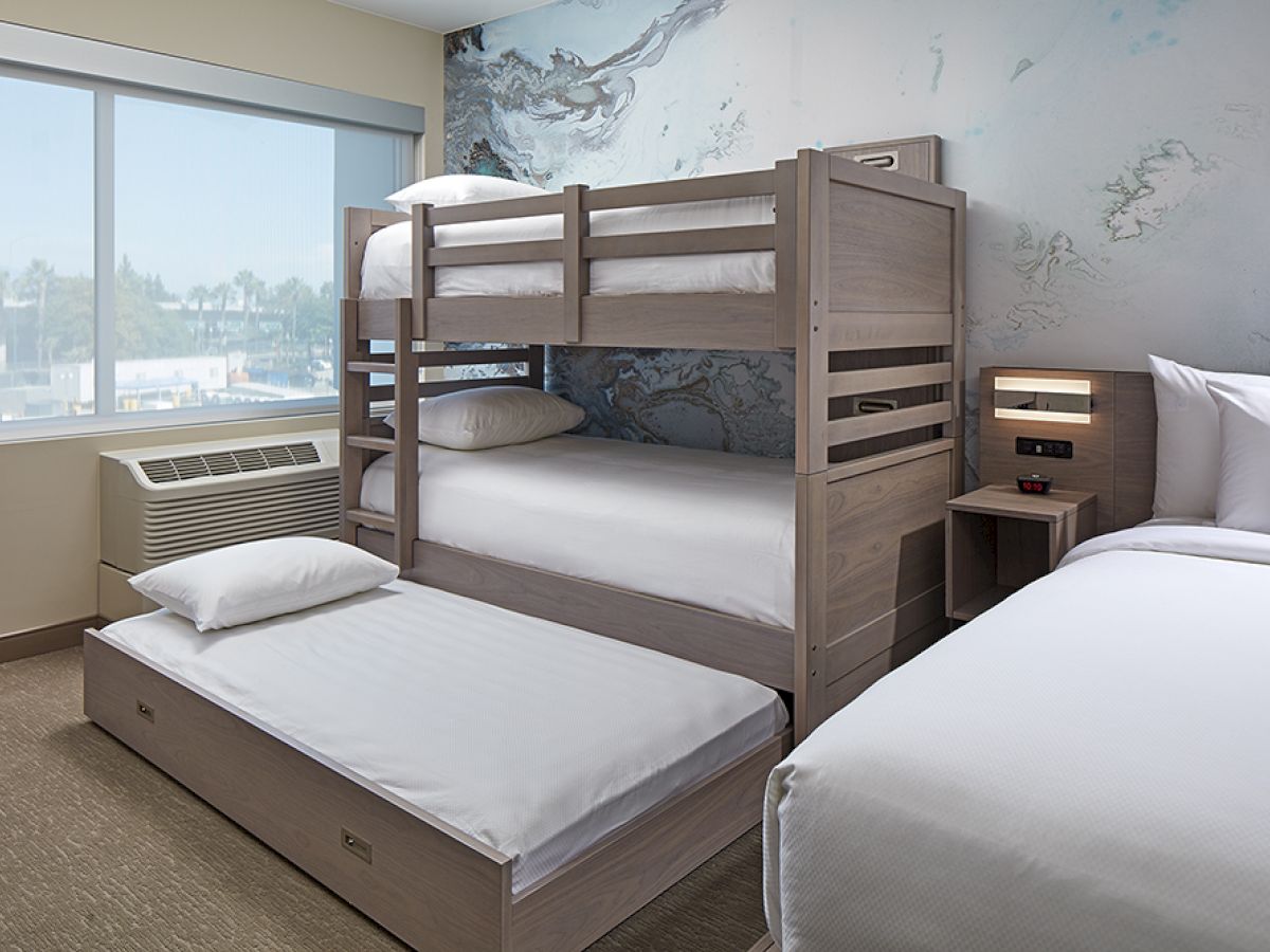 The image shows a hotel room with a bunk bed and a pull-out trundle bed beside a regular bed, along with a window, nightstand, and air conditioning unit.