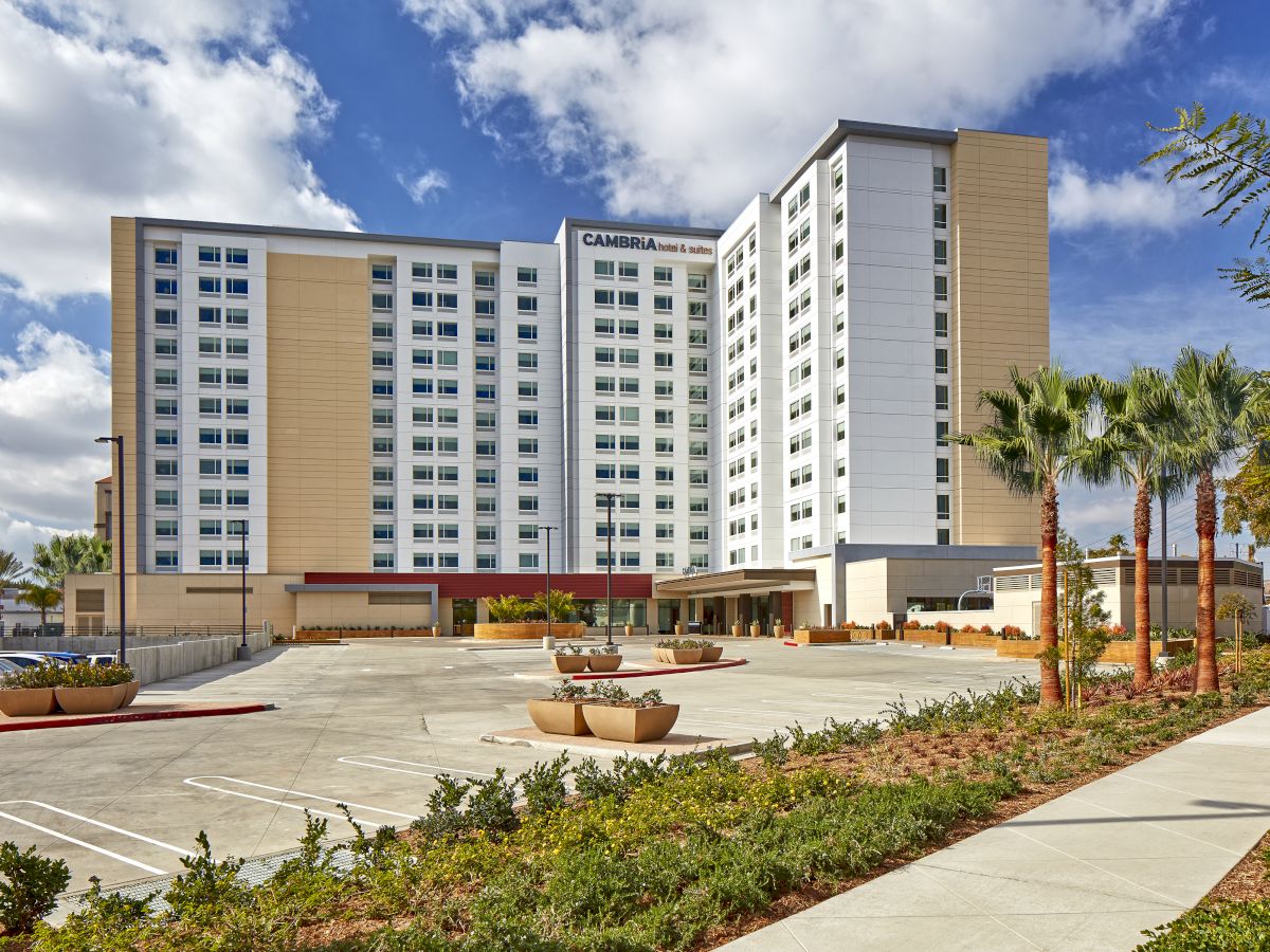 The image shows a multi-story building with a large, empty parking lot and palm trees in the foreground, under a partly cloudy sky.