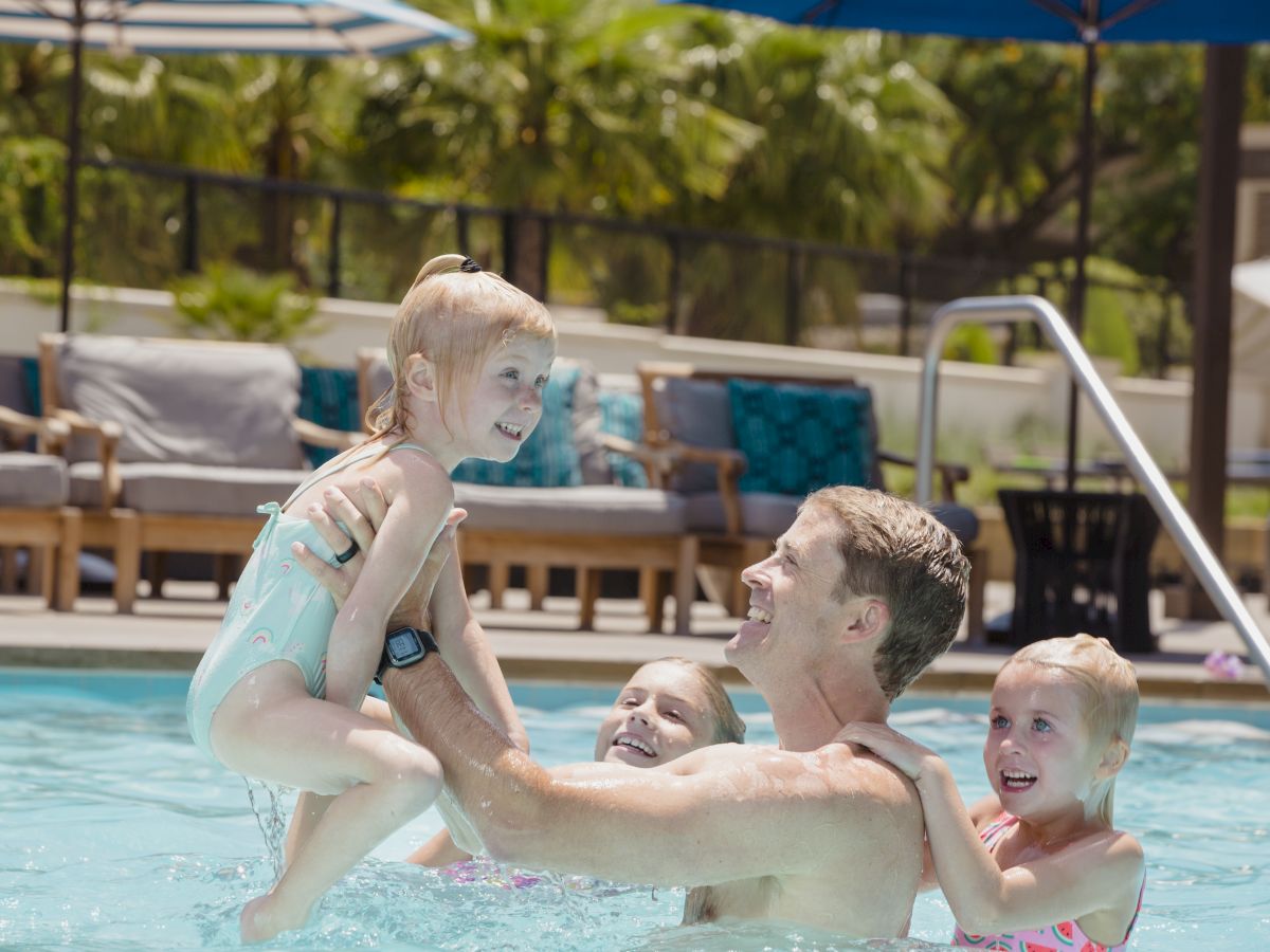 A man is playing in a pool with three young children, lifting one of them in the air. They all appear happy.