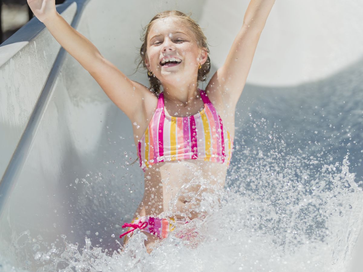 A child wearing a striped swimsuit is joyfully sliding down a water slide with arms raised, splashing as they reach the bottom.