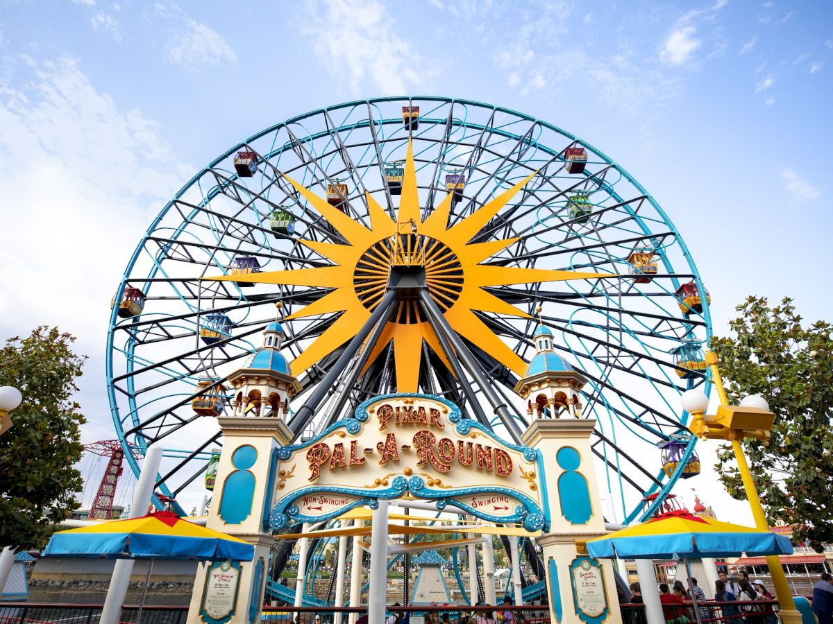 The image shows a large Ferris wheel with colorful cabins and a sun design at the center, located at a theme park on a bright day.