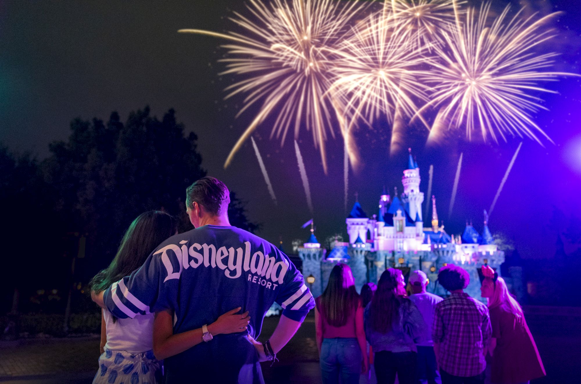 A couple and a group of people watch fireworks over a castle at Disneyland at night, creating a magical and joyful atmosphere.