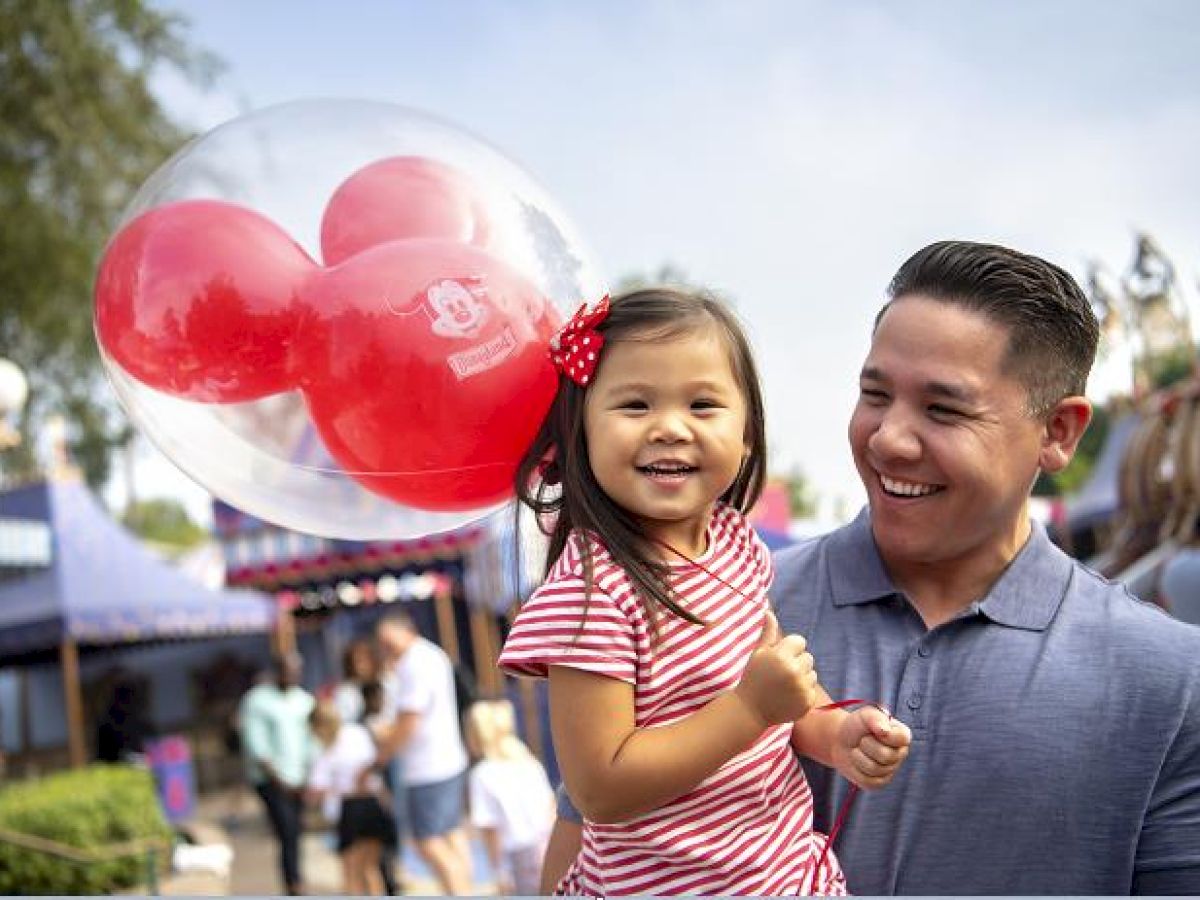 A man holds a smiling child with a Mickey Mouse balloon at an amusement park, surrounded by people on a sunny day.