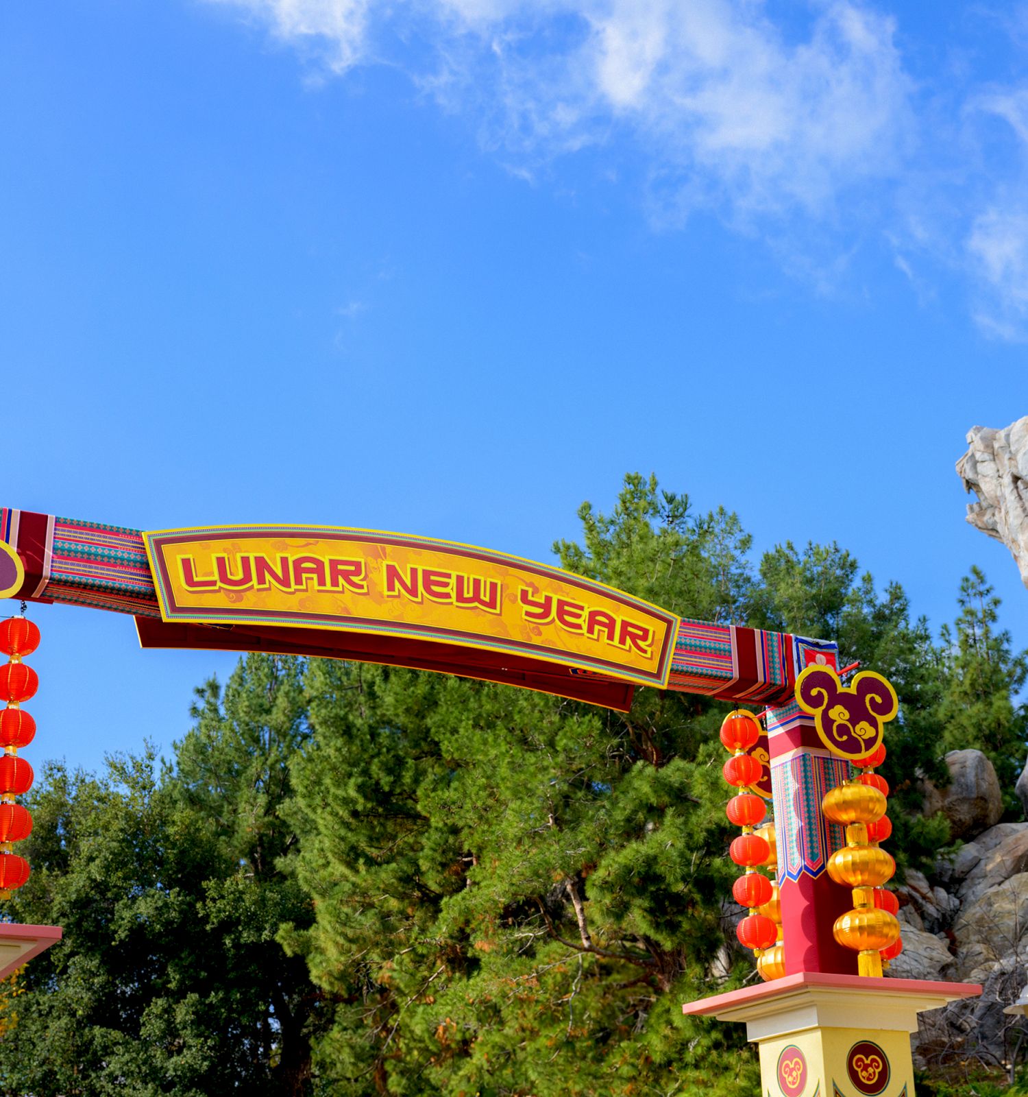 Lunar New Year sign with red lantern decorations set against a backdrop of trees and a rocky landscape under a clear blue sky.