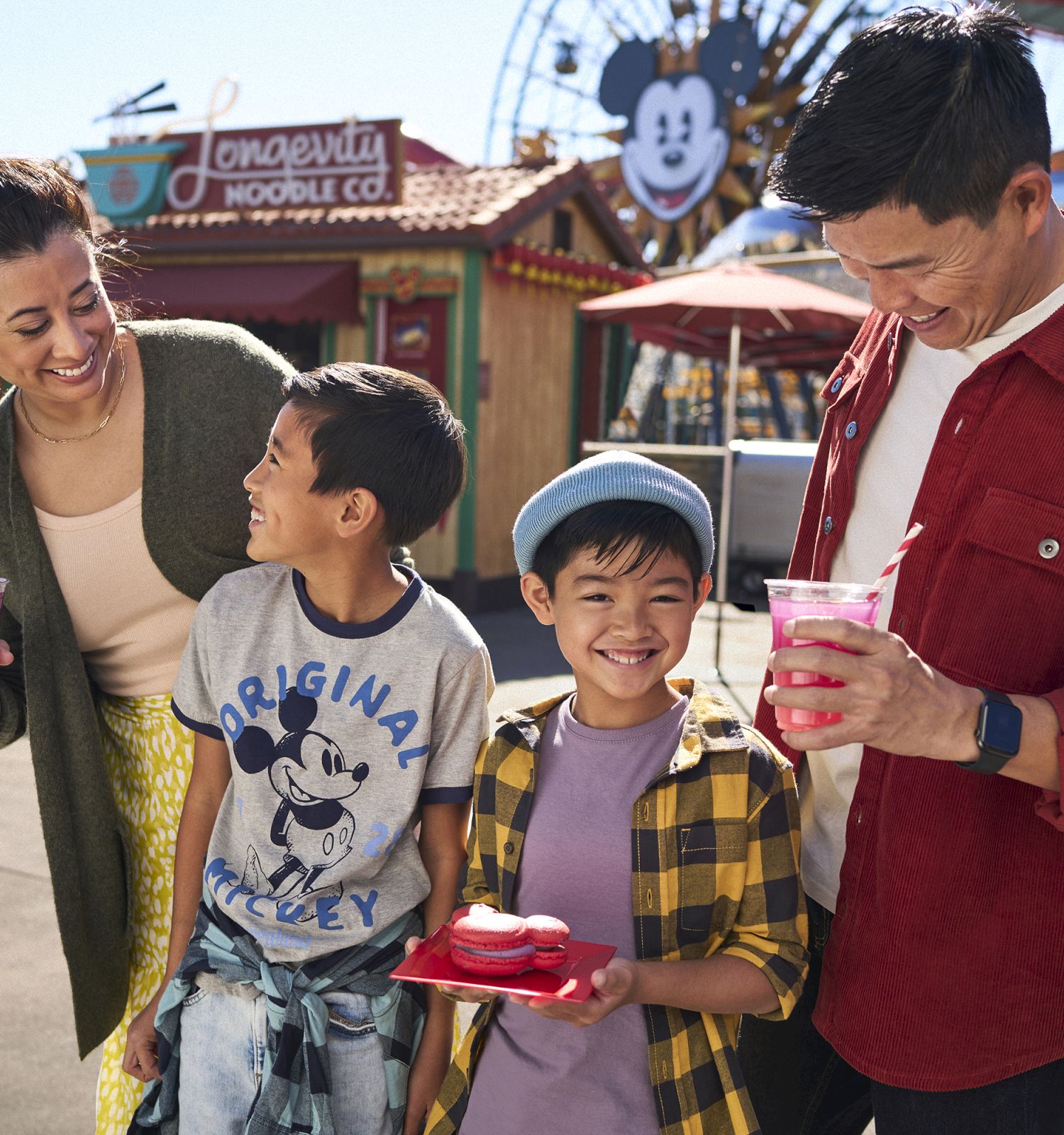 A family enjoying drinks and snacks at a theme park, with Mickey Mouse signage in the background. Smiling and having fun together.