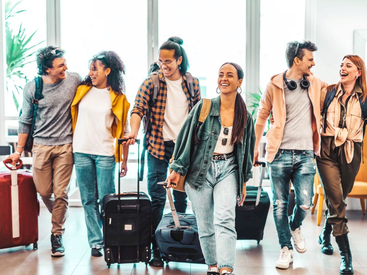 A group of six friends happily walk through an airport with their luggage and backpacks, chatting and smiling.