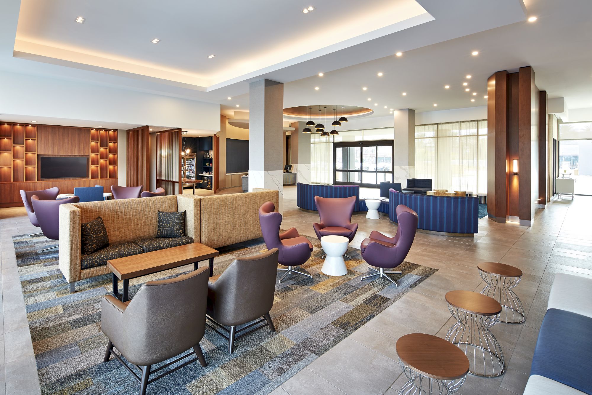 A modern, spacious hotel lobby with a mix of seating, including sofas, armchairs, and stools, featuring warm lighting and contemporary decor.