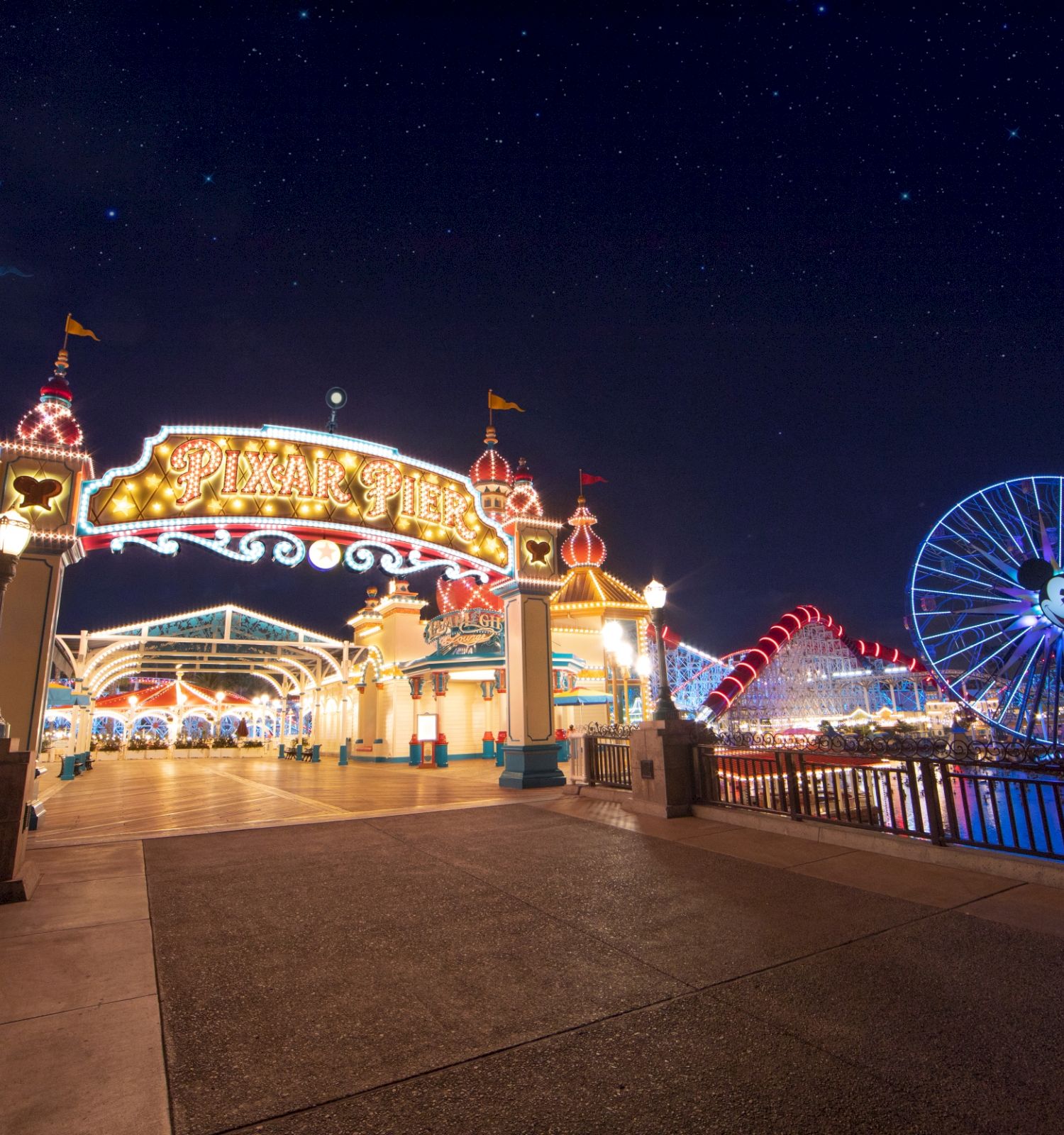 The image shows a brightly lit amusement park at night, featuring a sign for "Pixar Pier" and a large Ferris wheel in the background, illuminated with lights.