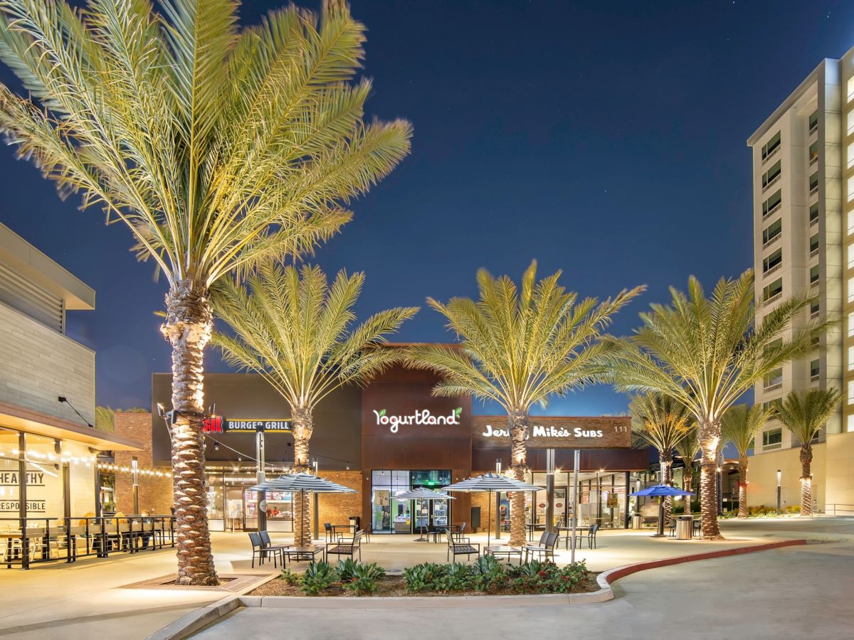 The image shows a well-lit outdoor shopping area with palm trees, featuring a Yogurtland and other stores around a seating area, at night.